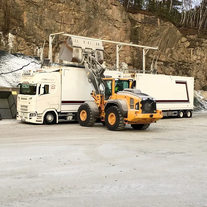 Scania-lastebil lastes av gul gravemaskin nær en fjellvegg.